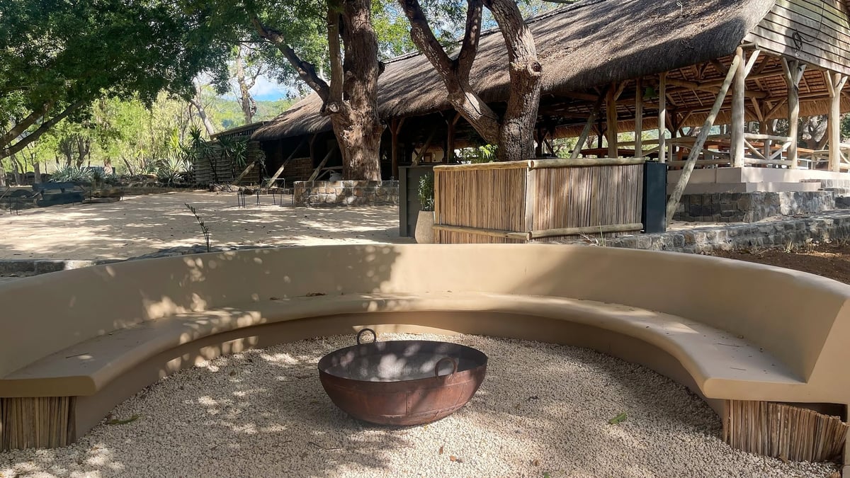 Curved tan concrete bench surrounds a metal fire pit on gravel base traditional thatched roof lodge under large trees