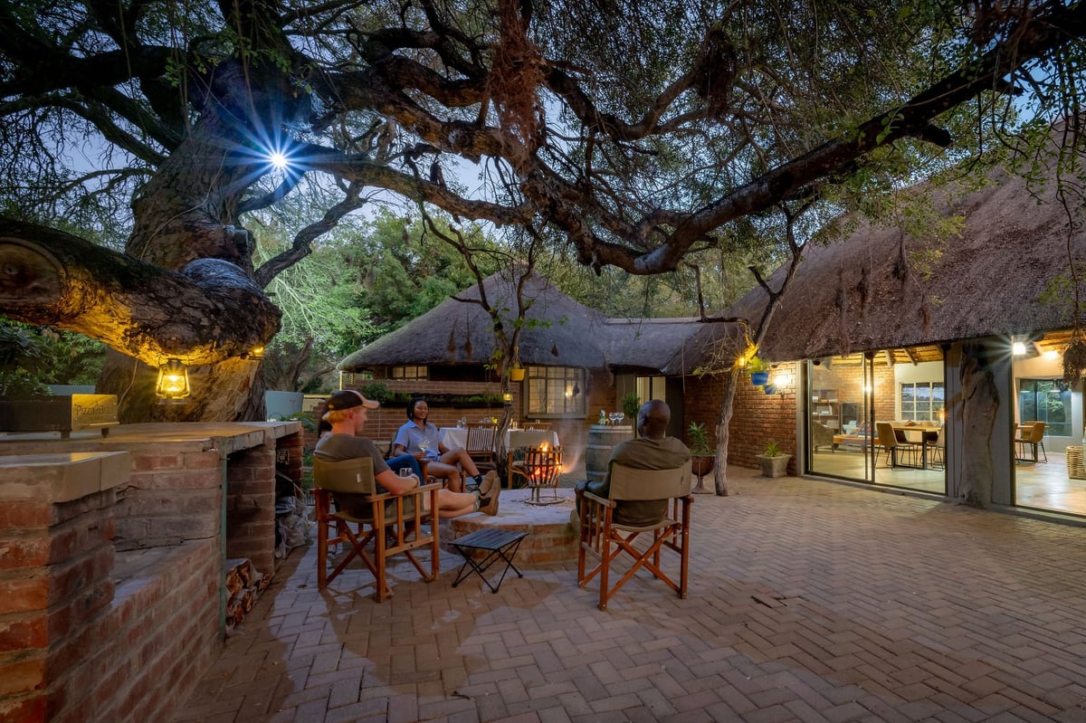 Evening outdoor courtyard under large tree with guests seated at tables near illuminated thatched-roof buildings and fireplace