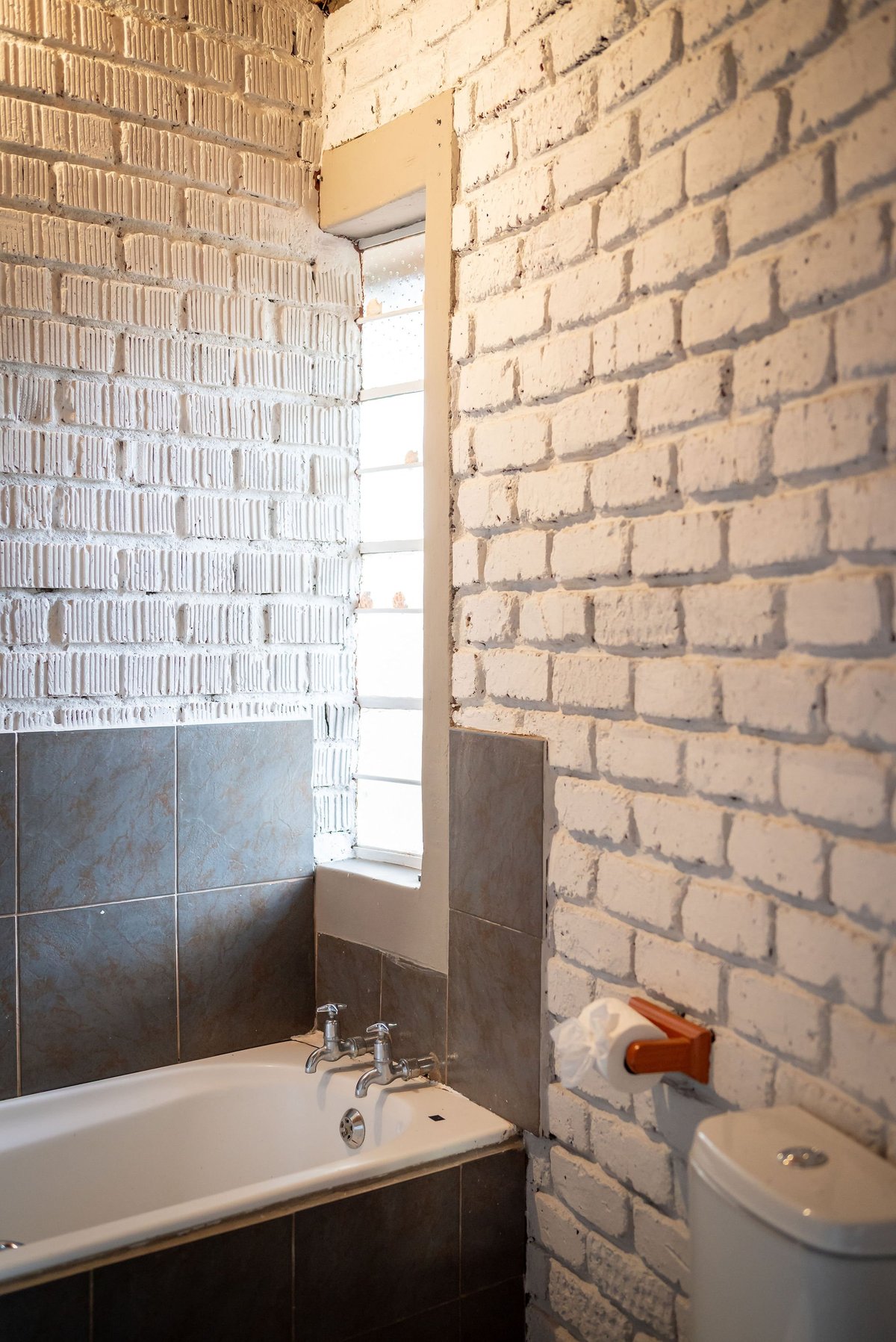 Small bathroom with white patterned tile wall, exposed brick wall, bathtub with fixtures, window, and toilet