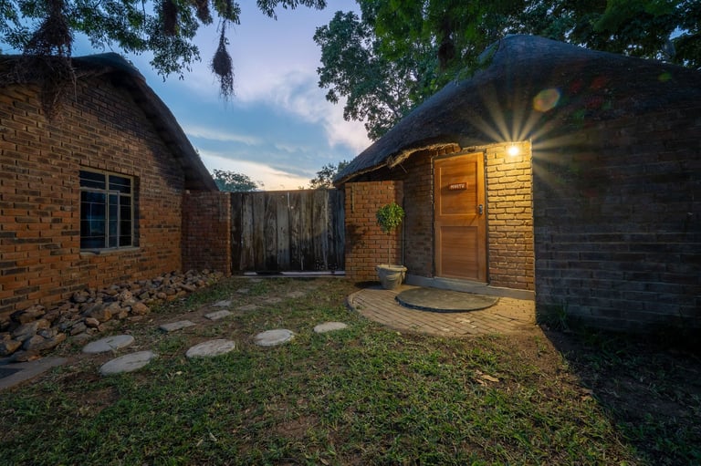 Brick courtyard at dusk with outdoor lamp, two residential buildings, stone pathway, and green lawn at twilight