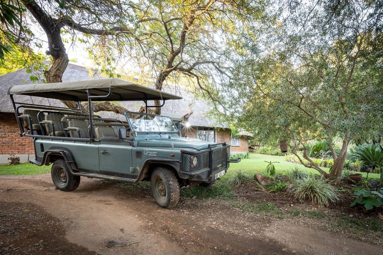 Blue Land Rover Defender parked under large trees with safari roof rack in a garden setting