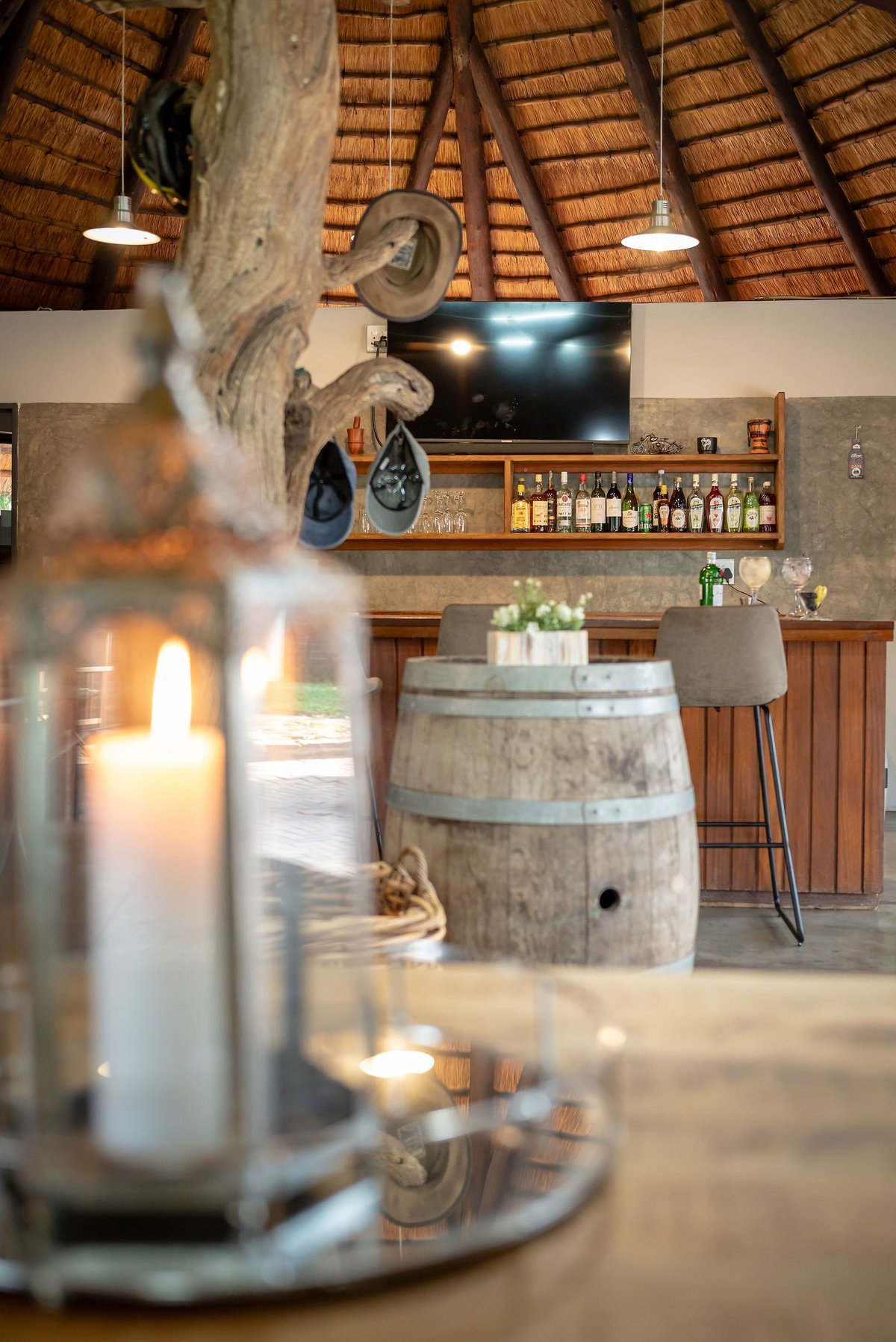 Interior of rustic bar with wooden barrel furniture, thatch roof, candles in foreground, and shelves of bottles behind counter