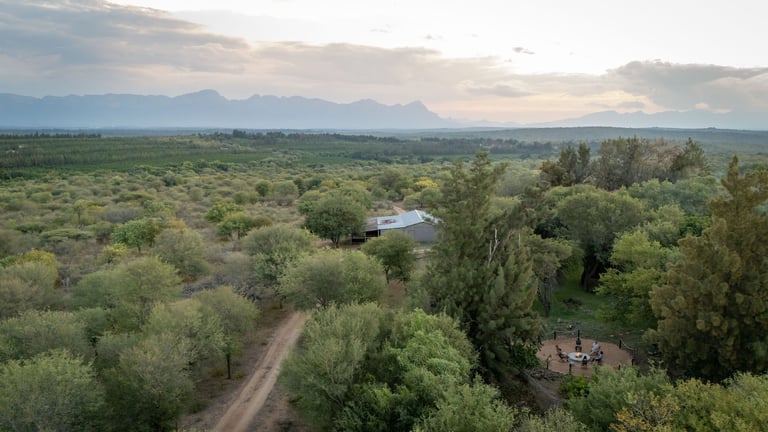 Aerial view of a rural landscape with dirt road, scattered buildings, trees, and mountains in the distance under cloudy sky