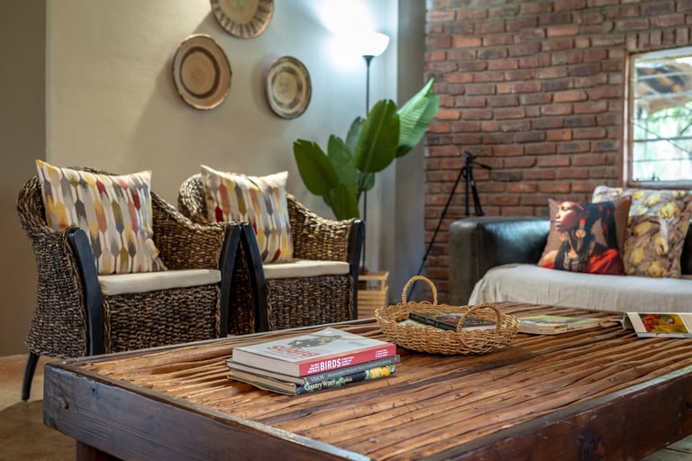 Rustic living room with wooden coffee table, wicker armchairs with patterned pillows, exposed brick wall, and decorative hats above seating