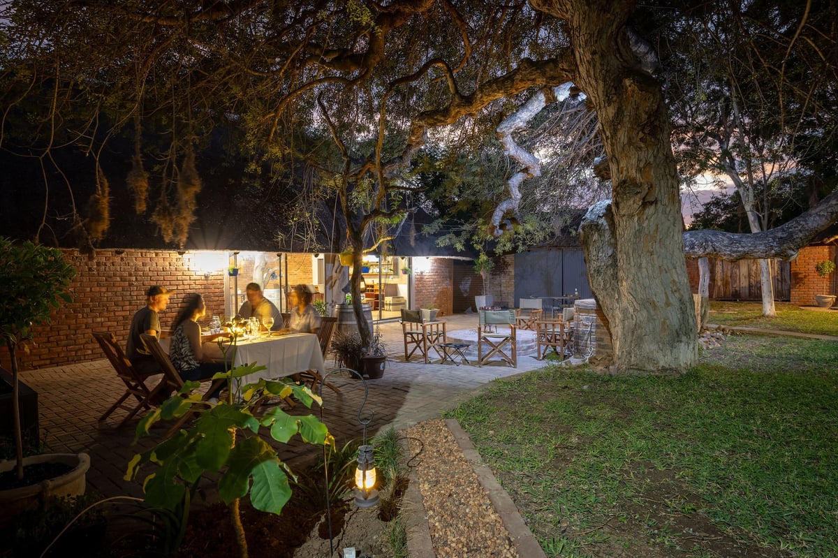 Evening backyard with illuminated brick house, large trees, green lawn, and outdoor seating areas