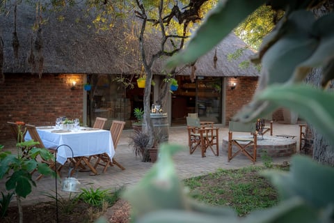 Outdoor courtyard dining area with white-clothed table, wooden chairs, brick walls, and trees, viewed through green foliage
