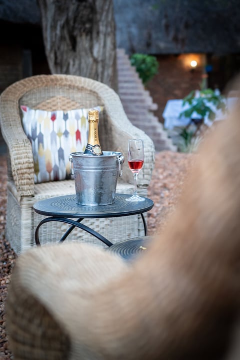 Champagne bottle in ice bucket on table beside wicker chair with patterned pillow in courtyard setting at dusk