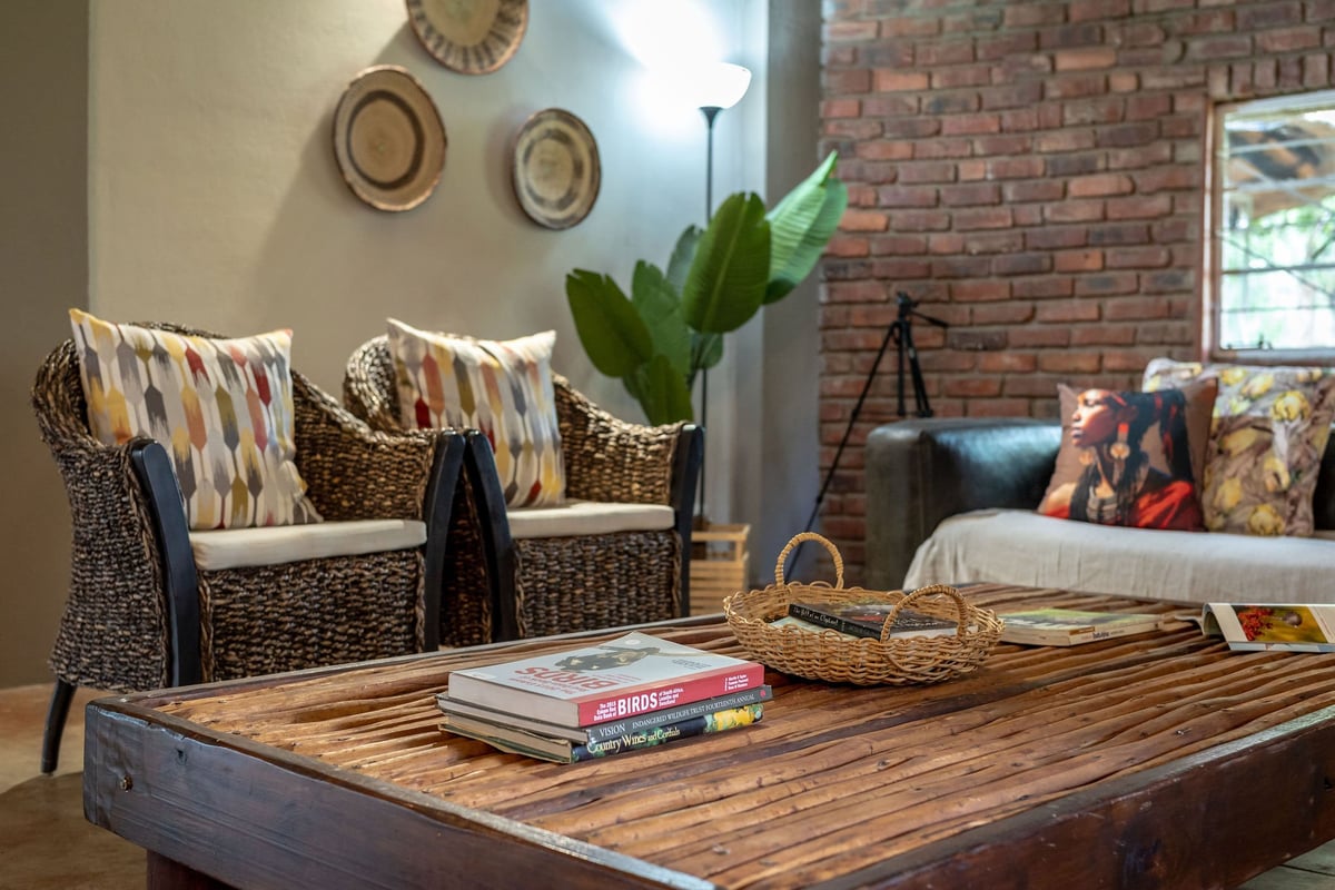 Living room with woven armchairs, wooden table with books, brick wall, and decorative hats on beige wall