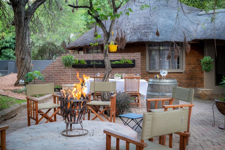 Outdoor patio with fire pit surrounded by wooden chairs, brick cottage, and large shade trees in background