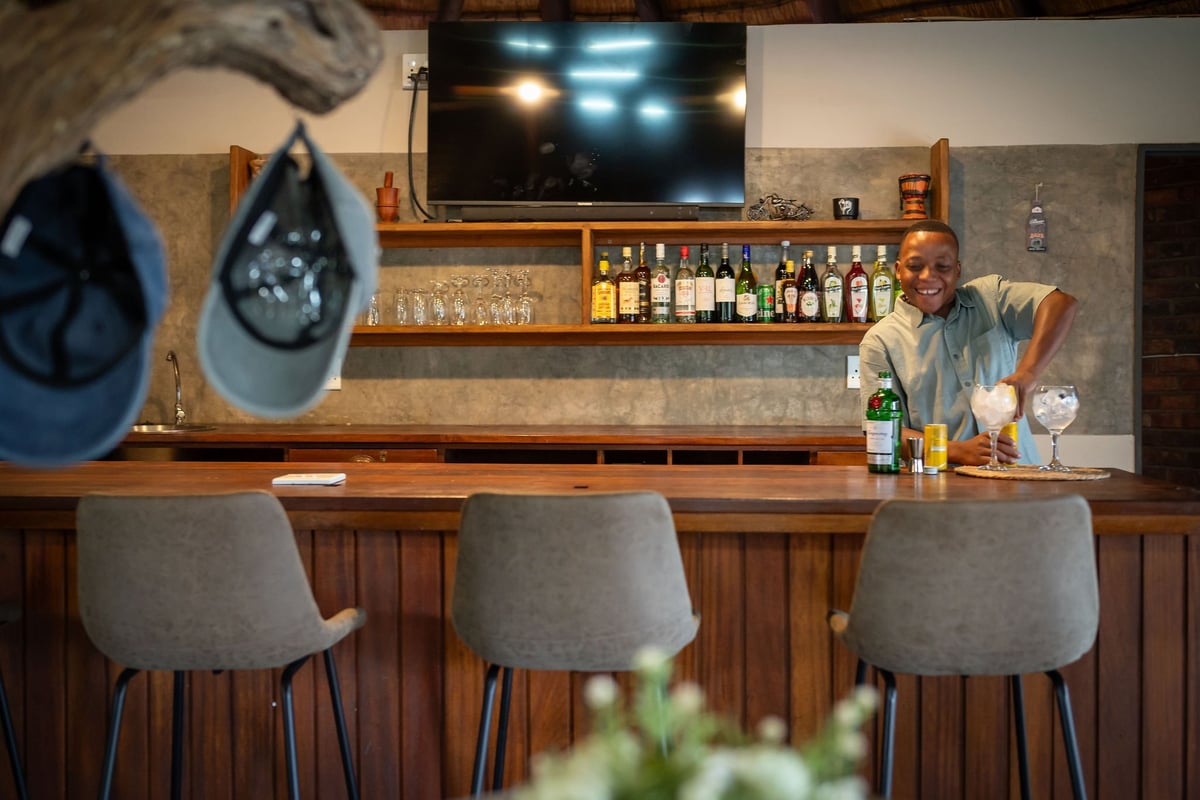 A well-stocked bar with three gray upholstered bar stools, wooden counter, shelves of liquor bottles, wall-mounted TV, and decorative blue hats displayed above