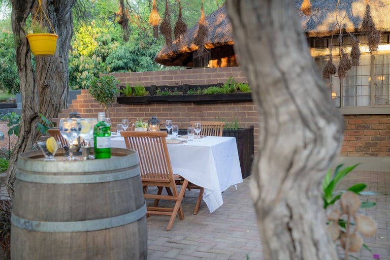 Outdoor dining setup in a brick courtyard with wooden chairs, a white-topped table, large trees, and a stone building with windows in the background