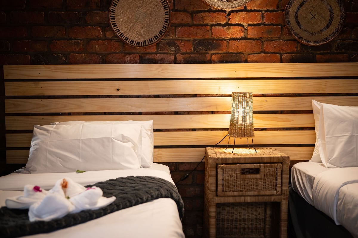 Rustic hotel bedroom with two twin beds, white pillows, wooden headboard, wicker nightstand, and clocks on exposed brick wall