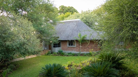 Brick cottage with thatched roof surrounded by lush green trees, tropical plants, and manicured lawn