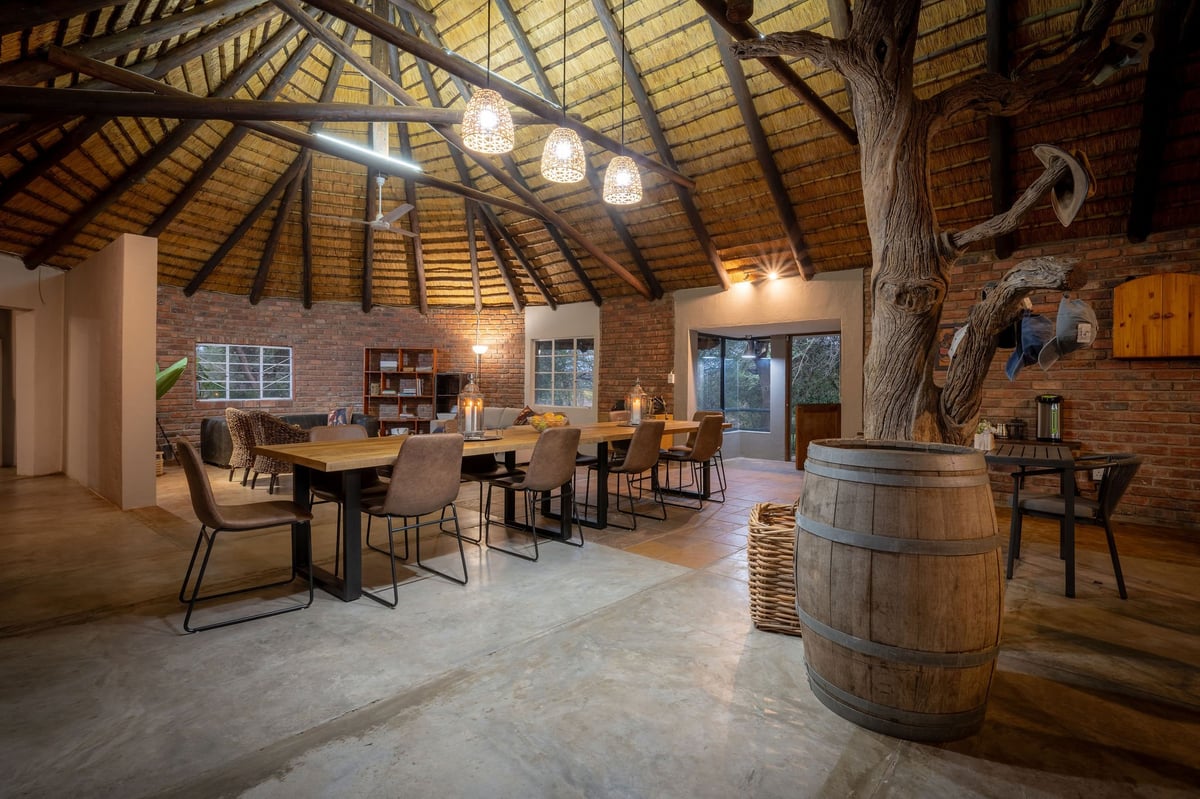 Modern rustic interior space with exposed wooden beams, dining tables, chairs, a large wooden barrel, and brick walls with ambient lighting