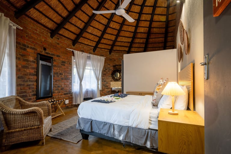 Spacious bedroom with exposed brick walls, wooden ceiling fan, white bed, wicker chair, and natural light from windows