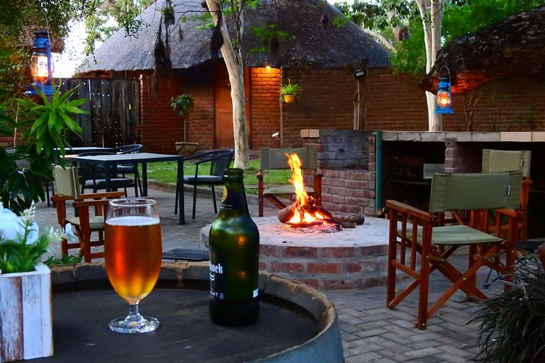 Outdoor lodge courtyard with firepit, beer glass and bottle on table, rustic wooden buildings with thatched roofs, and tropical plants