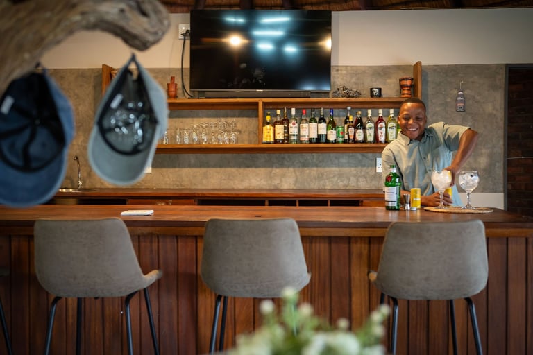 A modern bar interior with wooden counter, gray bar stools, mounted TV, shelves of liquor bottles, bartender in background, and decorative hats on wall