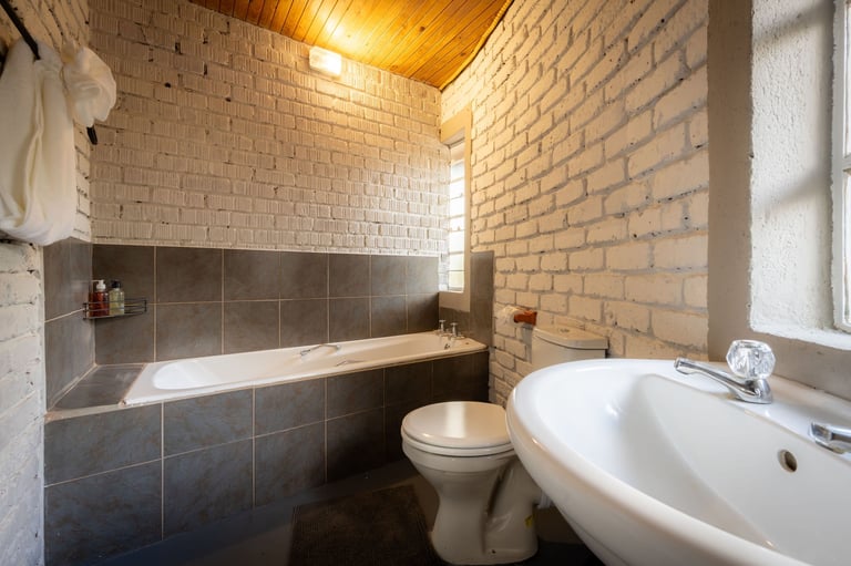 Modern bathroom with white pedestal sink, toilet, and bathtub with dark tile surround and exposed brick walls