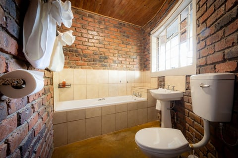 Rustic bathroom with exposed red brick walls, bathtub with cream tiles, pedestal sink, and wooden ceiling