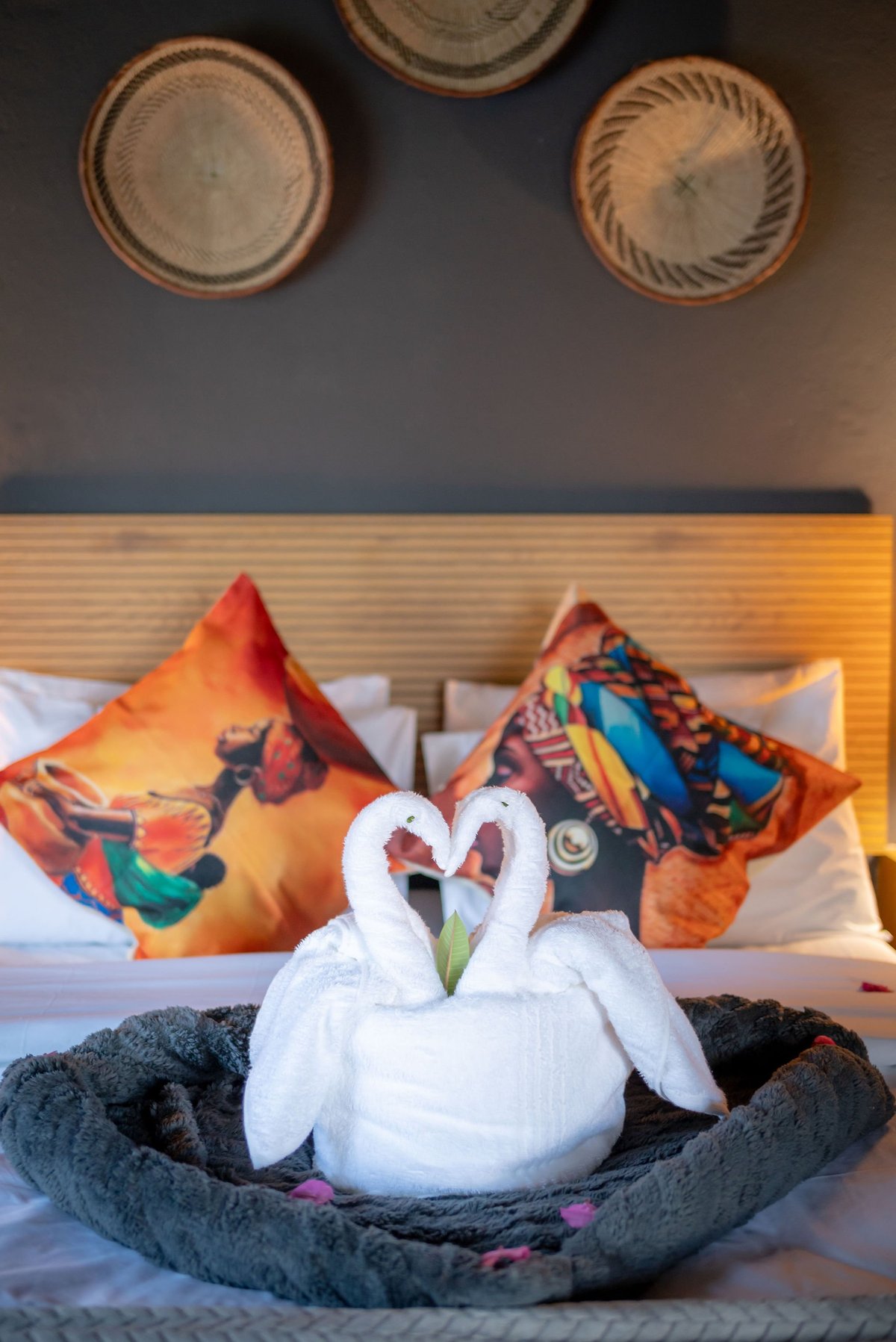 Two white towel swans form a heart shape on a bed with colorful decorative pillows and woven baskets on the wall above