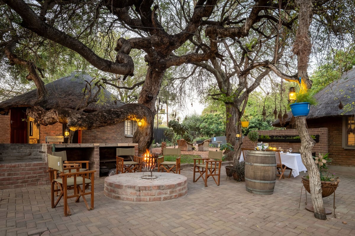 African safari lodge courtyard with thatched-roof bungalows, outdoor seating, fire pit, and large trees at sunset