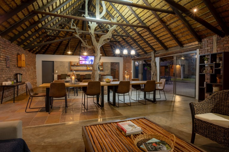 Modern open-plan indoor space with wooden beam ceiling, dining table with chairs, concrete flooring, and lounge seating area