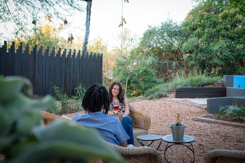 Two people sitting in a garden courtyard with potted plants, wooden fence, and gravel ground on a sunny day