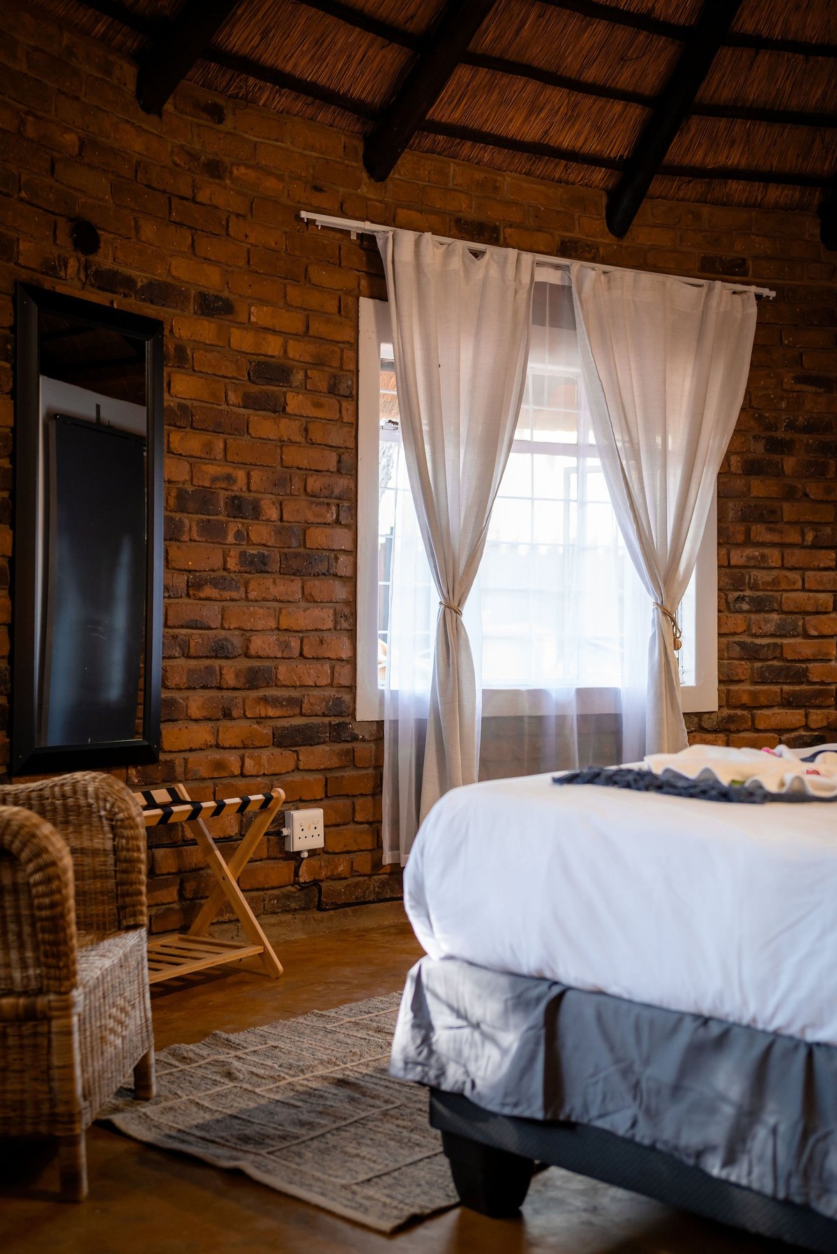 Rustic bedroom with exposed brick walls, wooden beam ceiling, bed with white bedding, cream curtains on window, and wooden luggage rack