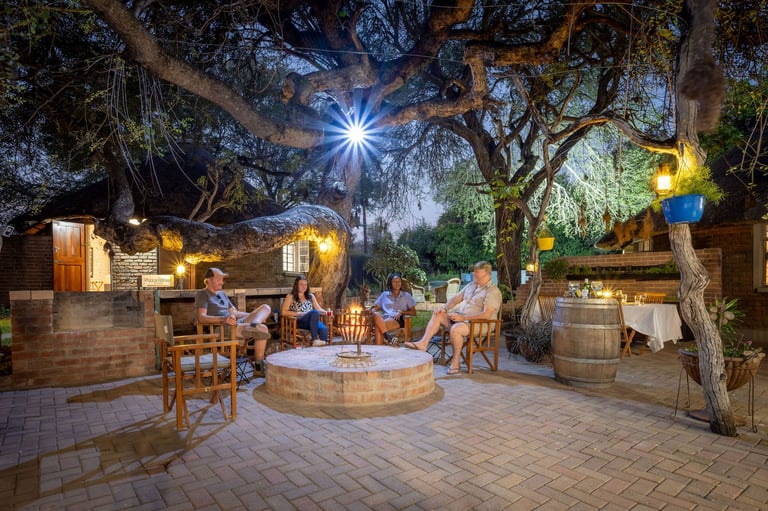 Outdoor courtyard at dusk with people gathered around a central fire pit, illuminated by string lights and trees overhead