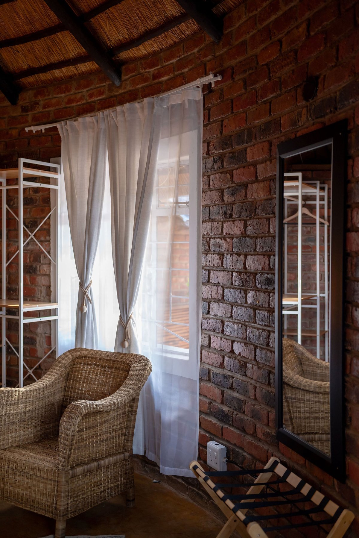 Rustic brick interior with wicker armchair, sheer curtains on window, and wooden ceiling beams