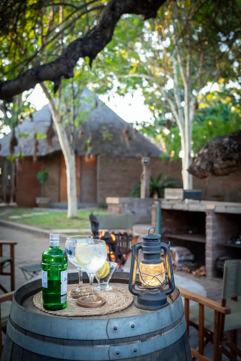 Barrel table with drinks and lantern in a tree-shaded courtyard with outdoor fireplace in background