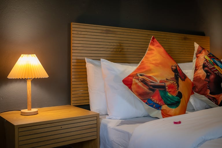 Modern bedroom with wooden headboard, white bedding, colorful decorative pillows, table lamp with yellow shade, and warm lighting