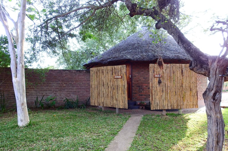 Traditional thatched-roof hut with wooden doors in a grassy yard surrounded by a brick wall and large trees