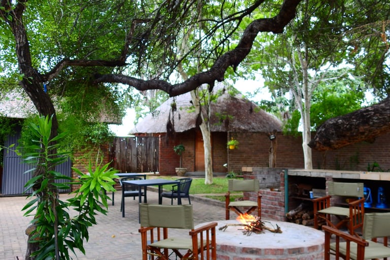 Outdoor courtyard with wooden tables, chairs, fire pit, and rustic huts surrounded by lush green trees