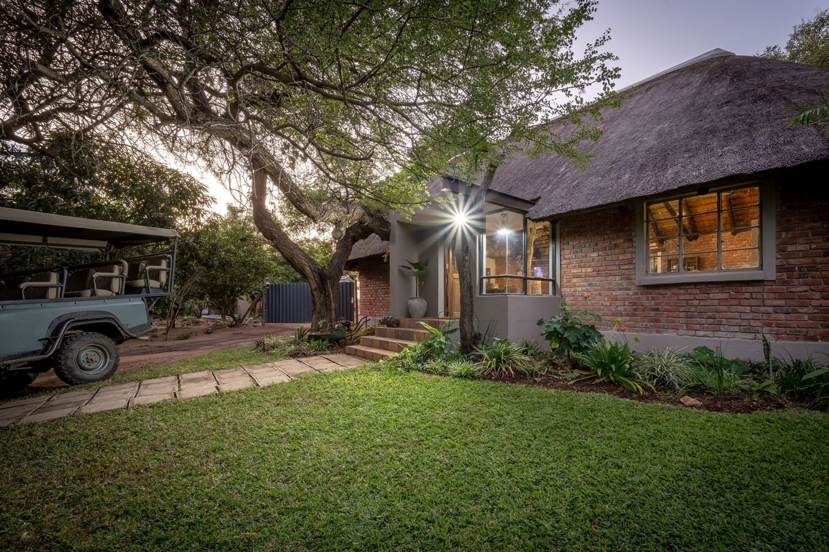 Modern brick house at dusk with thatched roof, illuminated entrance, manicured lawn, and parked vehicle under large tree