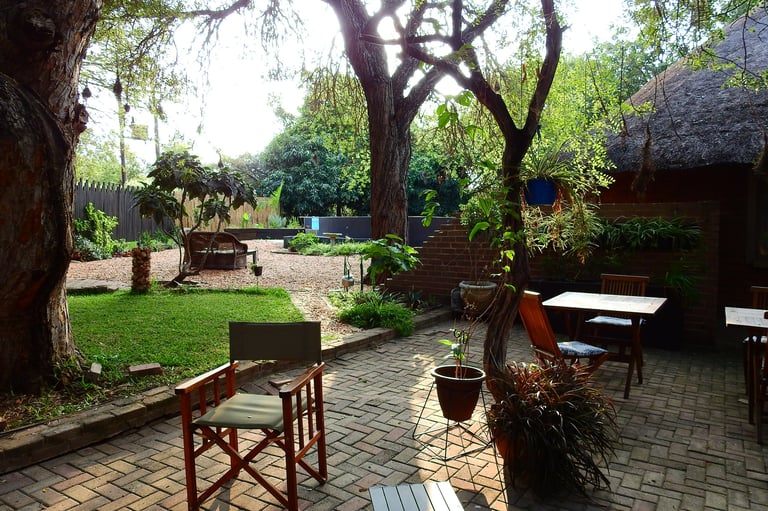Outdoor courtyard with brick patio, wooden chairs, and mature trees providing shade over green grass and rustic structures