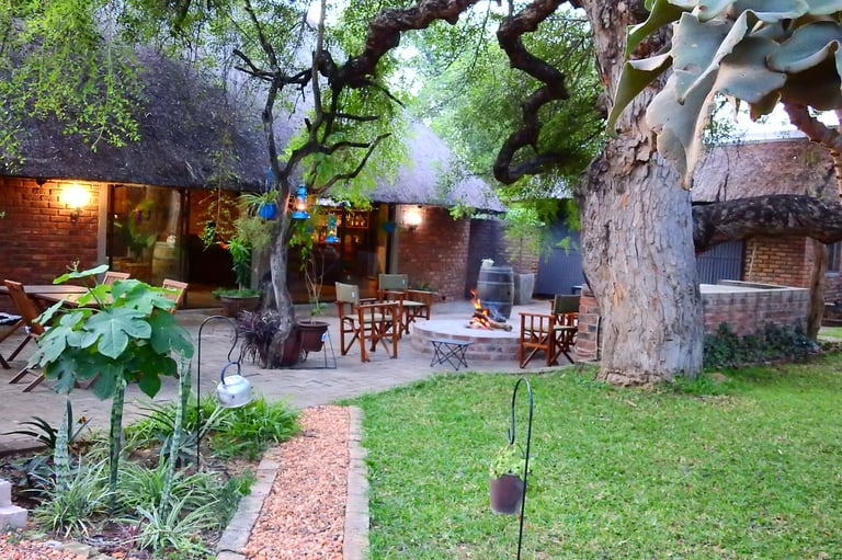 Rustic courtyard with large ancient trees, brick buildings, wooden chairs, and green lawn surrounded by natural landscaping at dusk