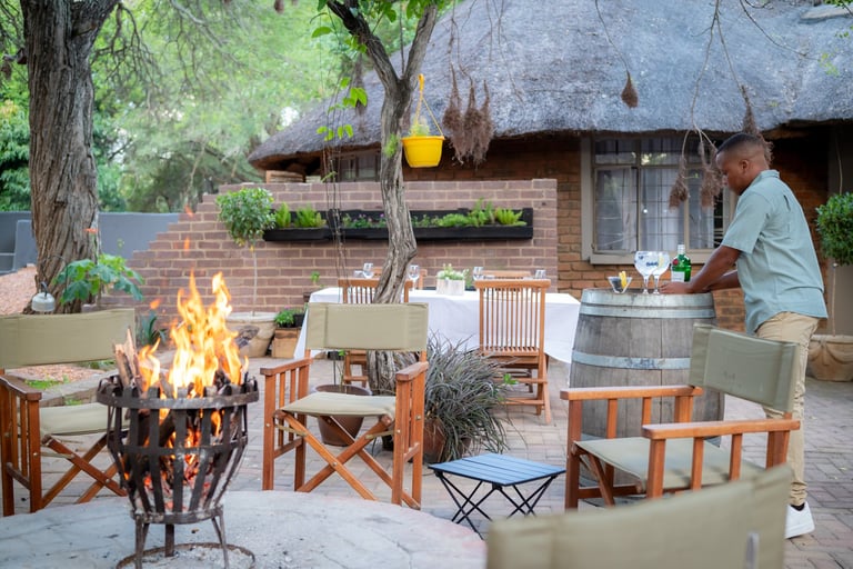 Outdoor safari lodge setting with fire pit, wooden chairs, and a man preparing food near a large barrel, with thatched-roof buildings in background