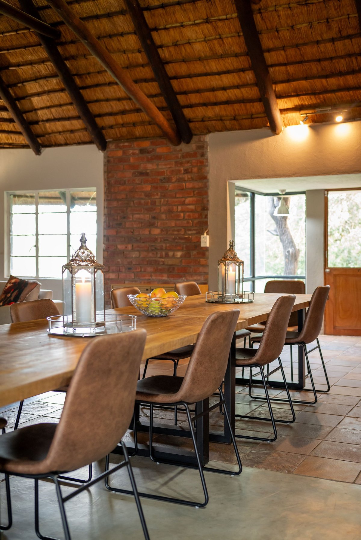 Modern dining room with wooden table, tan upholstered chairs, exposed brick chimney, wooden beam ceiling, and natural light from glass doors