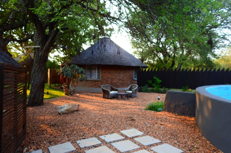 Brick cottage with thatched roof surrounded by green trees, patio seating area, and swimming pool in landscaped garden