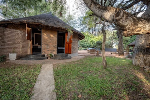 Brick cottage with thatched roof and red doors, surrounded by mature trees and green lawn with concrete pathway