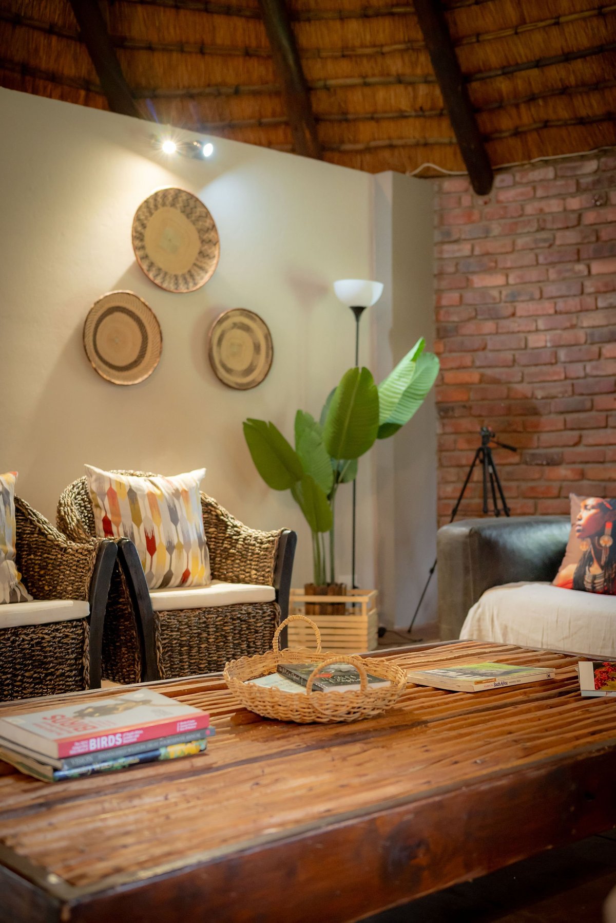 Warm living room with wooden table, wicker chair, decorative wall plates, potted plant, and exposed brick wall with rattan ceiling