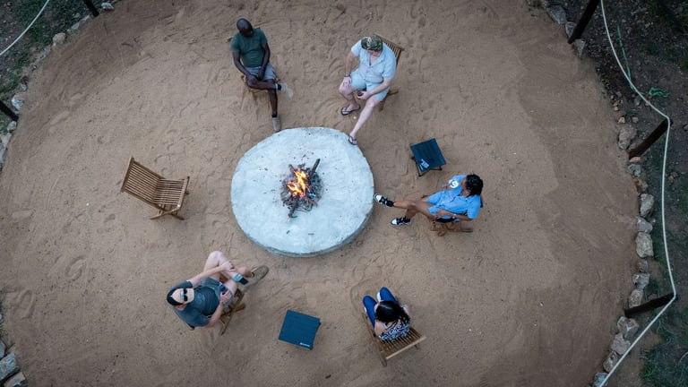 Aerial view of six people sitting around a white circular fire pit on sandy ground