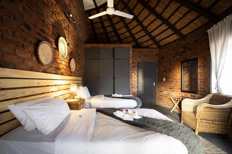 Modern bedroom with exposed red brick walls, wooden beam ceiling, white bedding, and woven chairs in a minimalist interior design