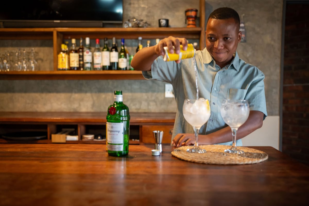 Bartender in blue shirt pouring drink into cocktail glass at wooden bar counter with bottle and bar setup in background