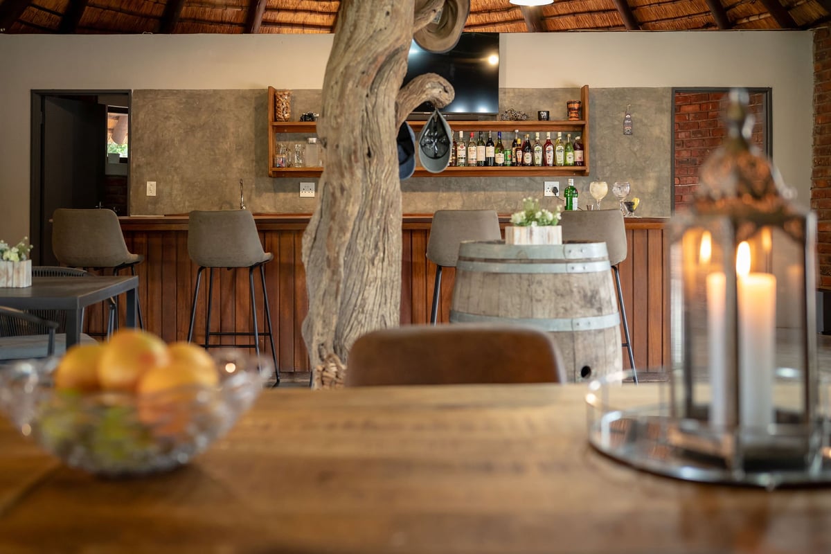 Interior of a rustic restaurant with a large wooden table in foreground, bar seating, exposed wooden beam, brick walls, and warm lighting