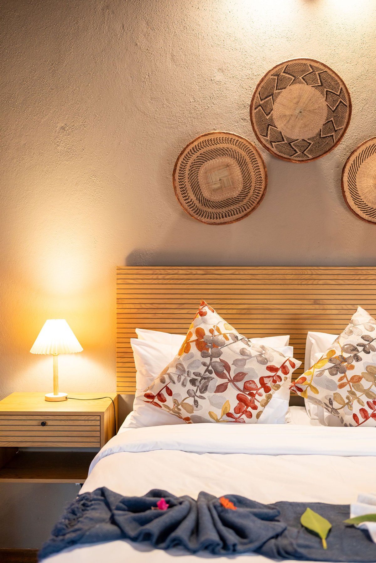 Cozy bedroom with wooden headboard, decorative woven plates on wall, lit bedside lamp, and white pillows with colorful accents