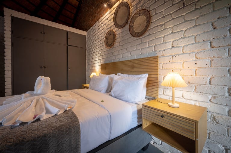 Modern bedroom with white brick walls, wooden headboard, neatly made bed with white bedding, nightstand with lamp, and decorative plates on wall