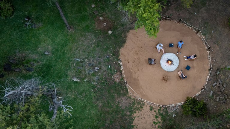 Aerial view of people gathered around a white circular shelter in a dirt clearing surrounded by green grass and trees