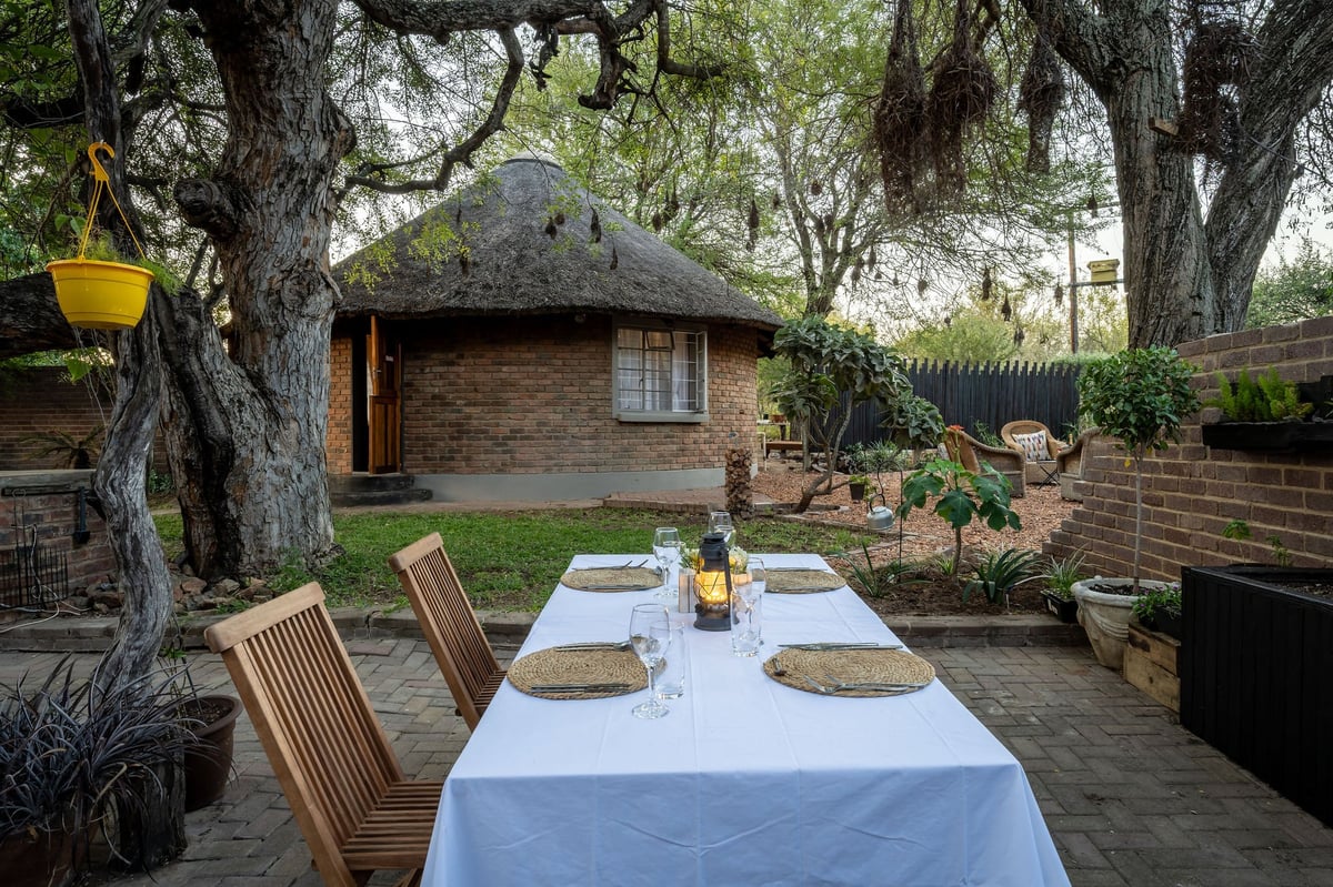 Outdoor dining setup with a table under large trees, facing a round brick cottage with a thatched roof